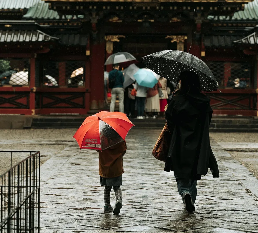 A rainy shrine courtyard with a crowd of strangers in the gateway and a metal railing intruding on the corner