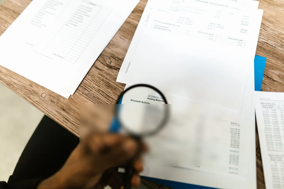 A hand holding a magnifying glass over a bank statement, with one section of account activity blurred out while the rest of the desk and paperwork stays untouched
